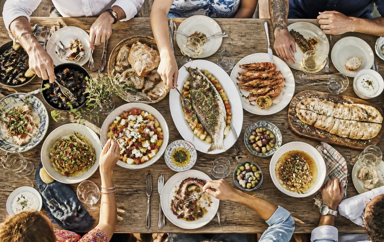 Overhead view of a long wooden table spread with local dishes, seafood, bread and wine, with guests reaching in to share the meal.
