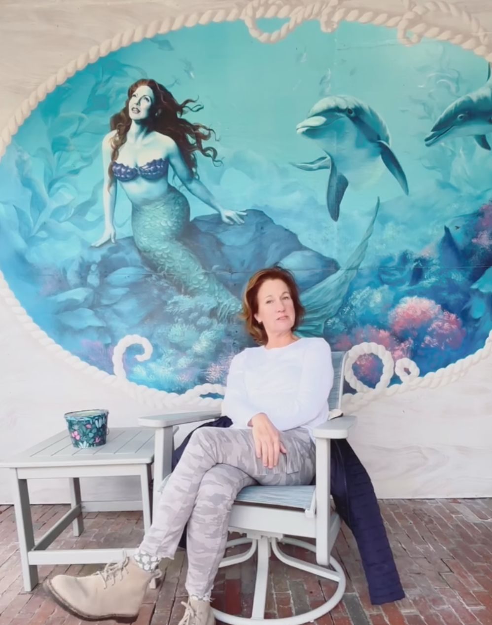 Katt seated in a chair in front of the painted mermaid and dolphin mural at one of The Rather Shacks properties
