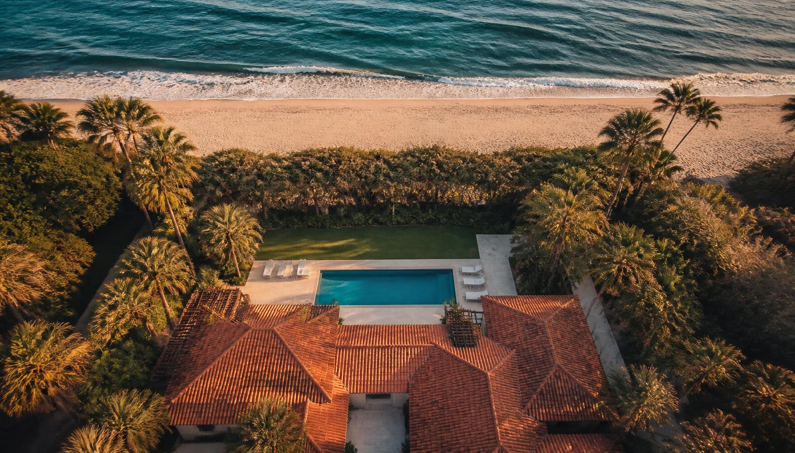 Aerial drone photograph of a coastal vacation rental home showing its pool, garden, tree line, the beach and the ocean beyond.